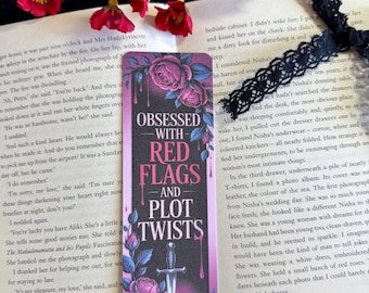 May include: A bookmark with the text "Obsessed with Red Flags and Plot Twists" and a dagger illustration, surrounded by pink roses and leaves. The bookmark is set on an open book with red flowers.