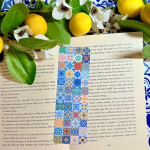 Peut inclure: Un marque-page coloré avec un motif de mosaïque de carreaux bleus, oranges et blancs. Il est posé sur une page de livre, entouré de citrons, de fleurs blanches et de feuilles vertes.