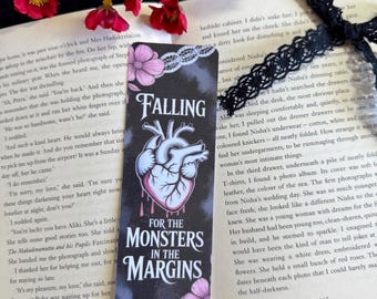 May include: A black bookmark with the text "FALLING FOR THE MONSTERS IN THE MARGINS" and a graphic of a heart. The bookmark is decorated with pink flowers and white lace. Red flowers and a book are in the background.
