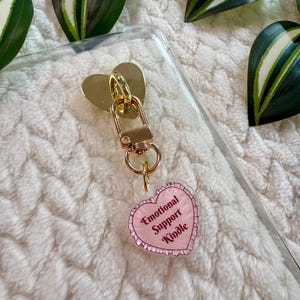 Op de afbeelding: Een goudkleurige hartvormige sleutelhanger met een sluiting en een kleine, roze hartvormige bedel. De bedel heeft de woorden "Emotional Support Kindle" in een decoratief lettertype. De achtergrond is een zacht, wit, getextureerd oppervlak met groen-witte bladeren.