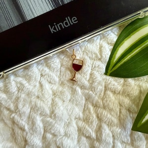 Könnte beinhalten: Ein goldfarbener Weinglas-Anhänger mit roter Weinfüllung. Der Anhänger ist an einem durchsichtigen Plastik-Lesezeichen befestigt. Das Lesezeichen ist an einem schwarzen Kindle E-Reader befestigt.