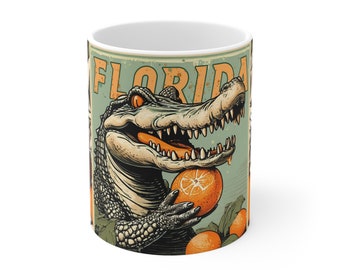 Puede incluir: Una taza de cerámica blanca con una ilustración de estilo vintage de un caimán comiendo una naranja. El texto "FLORIDA" está impreso en letras naranjas en la taza.