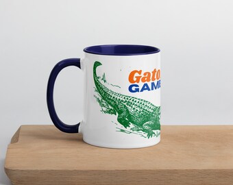 Puede incluir: Taza de cerámica blanca con borde azul. La taza presenta una ilustración de un cocodrilo verde y el texto "Gator Game" en naranja y azul.