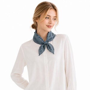 May include: A blue patterned neck scarf tied in a bow, worn over a white button-down shirt. The scarf has a delicate floral design and is made of a soft fabric. The scarf adds a touch of elegance to the outfit.