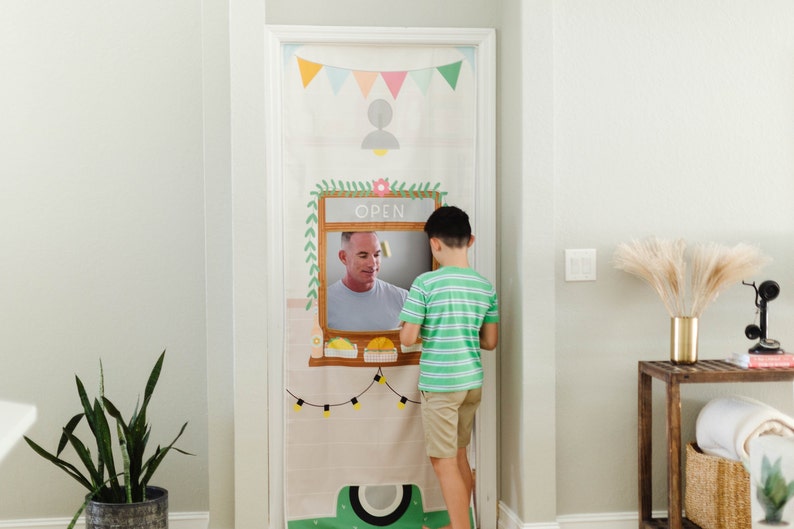 Puede incluir: Un ni&ntilde;o interact&uacute;a con una cubierta de puerta de puesto de comida de juguete. La cubierta de la puerta presenta la cara sonriente de un hombre en una ventana con la palabra "ABIERTO" encima. La cubierta de la puerta est&aacute; decorada con banderines coloridos, luces de cadena y un dise&ntilde;o verde y blanco.