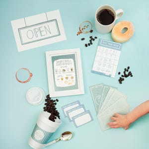 May include: An overhead shot of coffee-themed items on a light blue surface. Includes a white mug of coffee, a donut, coffee beans, a framed sign, a to-go cup, and order cards. The framed sign reads "Happy Brew Cafe & Bakery".