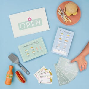 May include: A child's play food set with a hamburger, fries, and a hot dog. The set includes menus, play money, and an "OPEN" sign. The menu features tacos, burgers, and drinks. The set is on a light blue surface.