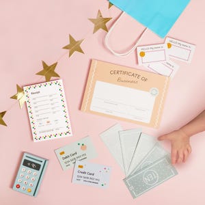 May include: A collection of toy business supplies, including a receipt, certificate, name tags, play money, debit and credit cards, and a calculator. The items are arranged on a pink surface with gold star decorations and a blue gift bag.