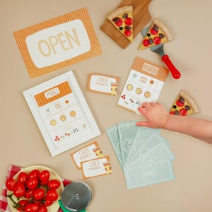 May include: A child's play pizza set with a menu, order card, play money, and pizza slices. The set includes a pizza cutter, a sign that says "OPEN", and name tags that say "CIAO! MY NAME IS".