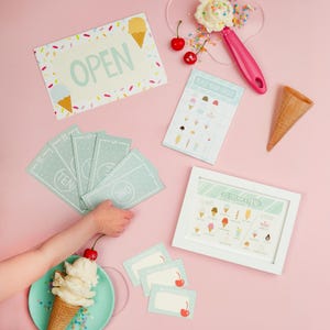May include: An assortment of ice cream-themed items on a pink background. Includes an "OPEN" sign, play money, a menu, order cards, and an ice cream cone. A scoop of ice cream with sprinkles and cherries is also present.
