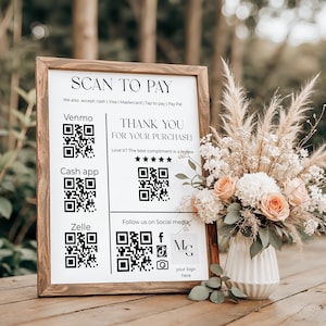 May include: A wooden framed sign with the text "SCAN TO PAY" and payment options. The sign also includes the text "THANK YOU FOR YOUR PURCHASE!" and social media icons. A floral arrangement with peach and white flowers sits beside the sign.