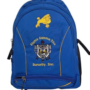 May include: Blue backpack with gold embroidered poodle and Sigma Gamma Rho Sorority, Inc. emblem. Features a zippered front pocket, black side pocket, and gold trim. The backpack has a top handle and adjustable shoulder straps.
