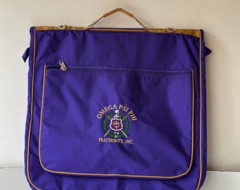 May include: Purple garment bag with a brown leather handle and trim. The bag has a zippered pocket and an embroidered logo that reads "Omega Psi Phi Fraternity, Inc."