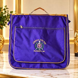 May include: A purple garment bag with gold trim and a top handle. The front features a zippered pocket and an embroidered Omega Psi Phi Fraternity, Inc. emblem in purple, gold, and green with crossed swords and a central shield design.
