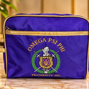 May include: A rectangular purple toiletry bag with gold trim and a side handle. The front features an embroidered Omega Psi Phi Fraternity, Inc. emblem with a crest, swords, and laurel wreath in gold, green, and white thread on the purple fabric.