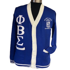 May include: A royal blue cardigan with white trim and buttons. The cardigan has a white embroidered Greek letter design on the left side and a white embroidered crest with the text "Phi Beta Sigma Fraternity, Inc." on the right side.