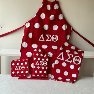 May include: Red and white polka dot kitchen set with Delta Sigma Theta sorority letters. The set includes an apron, two pot holders, and a dish towel.
