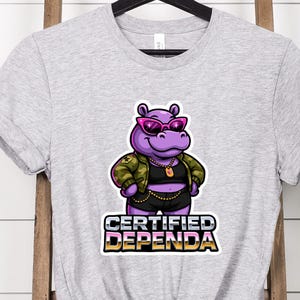 Op de afbeelding: Athletic heather grijze t-shirt met een cartoon nijlpaard met een zonnebril en een jas in militaire stijl. Het nijlpaard heeft een gouden ketting en de tekst "CERTIFIED DEPENDA" in een gele en paarse lettertype.