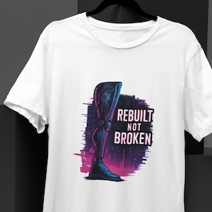 Può includere: T-shirt bianca con grafica di una gamba protesica futuristica nei toni del blu, viola e rosa. Il testo "REBUILT NOT BROKEN" è visualizzato sopra la gamba. Il design ha un'estetica cyberpunk.