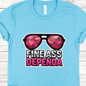 Op de afbeelding: Lichtblauw T-shirt met de tekst "FINE ASS DEPENDA" in roze en wit. De afbeelding bevat een zonnebril met roze camouflageglazen. Het shirt is gemaakt van een zacht materiaal.