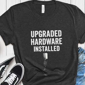 May include: Dark gray t-shirt with the text "UPGRADED HARDWARE INSTALLED" and a graphic of a computer component. The shirt is paired with black and white sneakers and denim shorts. A small potted plant is also in the image.
