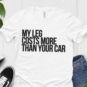 May include: White t-shirt with the text "MY LEG COSTS MORE THAN YOUR CAR" in black. The shirt is paired with black and white sneakers and blue denim shorts. Green tropical leaves are also in the image.