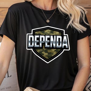 Può includere: T-shirt nera con la scritta "DEPENDA" in grassetto argentato, all'interno di un disegno a forma di scudo con un motivo mimetico. La maglietta ha maniche corte e girocollo.