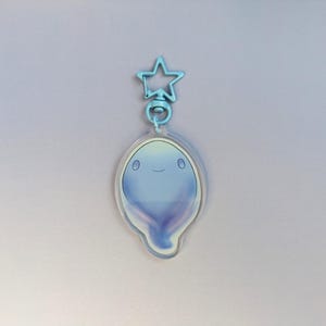 Puede incluir: Un llavero de acrílico transparente con un personaje azul sonriente con forma de fantasma. El llavero tiene un cierre en forma de estrella azul claro y un anillo plateado. El personaje tiene un degradado de colores azules y morados.