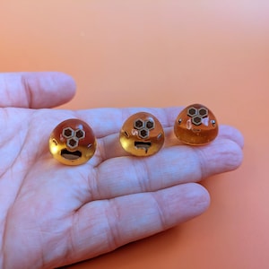 Könnte beinhalten: Drei bernsteinfarbene, kuppelförmige Zierknöpfe mit goldenem Sockel und geometrischem Design. Die Knöpfe werden auf einer Hand vor einem orangefarbenen Hintergrund präsentiert. Die Knöpfe haben ein einzigartiges, künstlerisches Design.