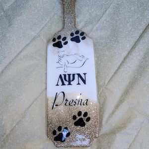 May include: A decorative paddle with a gold glitter handle and base. The paddle has a white center with black paw prints, a reclining cat illustration, and the Greek letters ΛΨΝ. The name "Dresha" is written in black script.