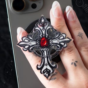 May include: A phone grip featuring a gothic-style cross design with intricate black and white filigree patterns and a central oval-shaped red gemstone. The accessory is attached to the back of a grey smartphone, providing a secure hold for the device.