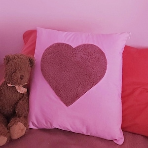 May include: A pink pillow with a heart-shaped patch of fuzzy pink fabric. A brown teddy bear sits on a pink and red bedspread.