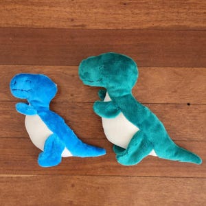 May include: Two plush dinosaur toys. One is bright blue, the other is teal. Both have white bellies and are lying on a wooden surface. These soft toys are designed to resemble a Tyrannosaurus Rex.
