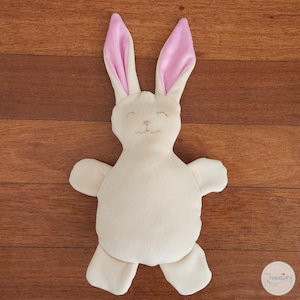 Puede incluir: Un juguete de peluche con forma de conejo de color crema, con el interior de las orejas rosa, rasgos faciales bordados y brazos y piernas extendidos. El conejo está sobre una superficie de madera. El juguete está diseñado para ser suave y tierno.