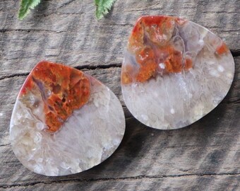 May include: Two teardrop-shaped agate cabochons with a white base and orange and red patterns. The cabochons are on a wooden surface.