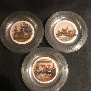 May include: Three pewter plates with a white porcelain center featuring images from the American Revolution. Each plate has the text "The Great American Revolution" and "1776" around the rim.