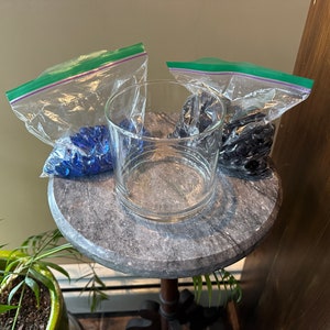 May include: A clear glass cylinder vase sits on a small, round, marble-topped table. Two plastic bags filled with blue glass beads and one with black glass beads are next to the vase. A green plant is in the foreground.