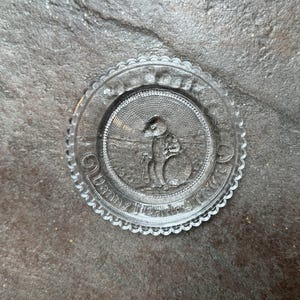 May include: A clear glass plate with a raised design of a person holding a ball. The plate has a scalloped edge and the words "Danny Meadow House" are inscribed around the edge.