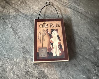 May include: A wooden sign with a red border and a brown background. The sign features a cartoon cat with black, white, and orange fur, sitting next to a potted plant. The text on the sign reads "Cats Rule!"