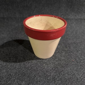 May include: A small, white ceramic planter with a red rim. The planter is empty and sitting on a dark gray surface.