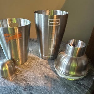 May include: Stainless steel cocktail shaker set with three pieces and a lid. One shaker has the word "COINTREAU" in orange, another has "COSMOPOLITAN" text. The set is designed for mixing drinks.