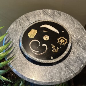 May include: A round, gray marble table top with a black circular inlay. The inlay has a rope border and features a compass, a treasure chest, a ship's wheel, an anchor, and a seashell.