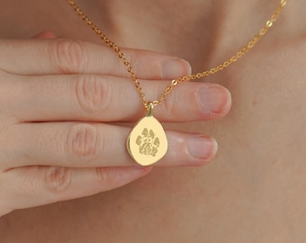 Può includere: Una collana color oro caratterizzata da una catenina delicata e un piccolo ciondolo dalla forma organica irregolare. Il ciondolo presenta al centro l'incisione dettagliata di un'impronta di zampa ed è appeso a una sottile catena in metallo.