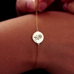 May include: A gold-colored bracelet featuring a circular pendant with a floral design and the word "August" engraved on it. The delicate chain is held by a hand against a dark background. The bracelet is a piece of jewelry.