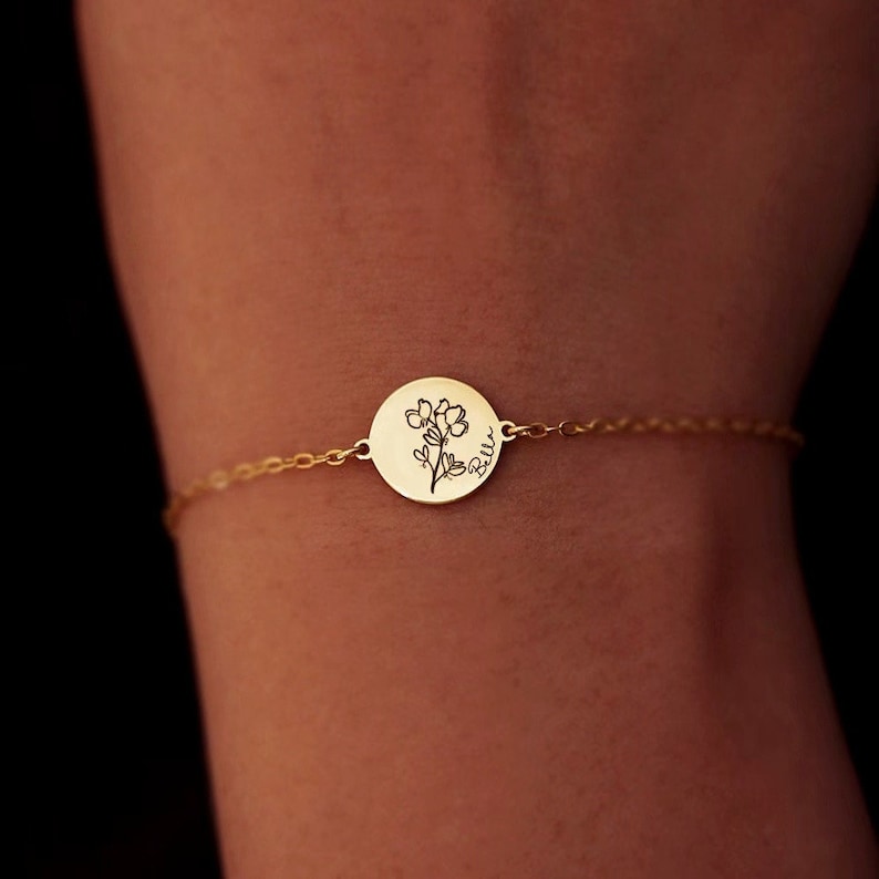 May include: A gold-colored bracelet with a delicate chain and a circular pendant. The pendant features a floral design and the name "Belle" in script. The bracelet is shown on a person's wrist.