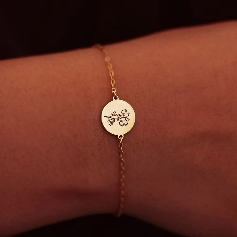 May include: A delicate gold-colored bracelet featuring a small, round charm with a floral design. The charm is connected to a thin chain. The bracelet is shown on a wrist, highlighting the jewelry's elegant simplicity.