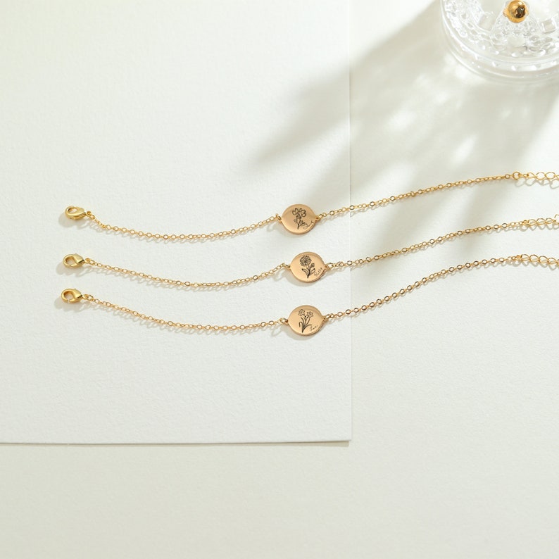 May include: Three gold-colored chain bracelets with oval pendants, each featuring a different floral design. The bracelets have a delicate chain and a lobster clasp closure. The pendants have engraved floral illustrations.