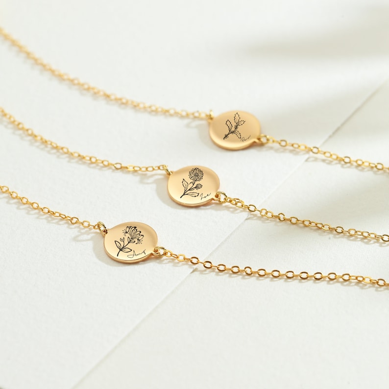 May include: Three gold-colored chain bracelets with circular pendants. Each pendant features a delicate floral illustration and a month name. The bracelets are displayed on a white surface, showcasing the intricate details of the jewelry.