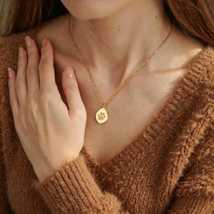 Puede incluir: Un collar de tono dorado con una cadena delicada y un peque&ntilde;o colgante de forma org&aacute;nica e irregular. El colgante presenta un sutil dise&ntilde;o de huella grabado en el centro, suspendido de una cadena de eslabones met&aacute;licos finos sobre el cuello.