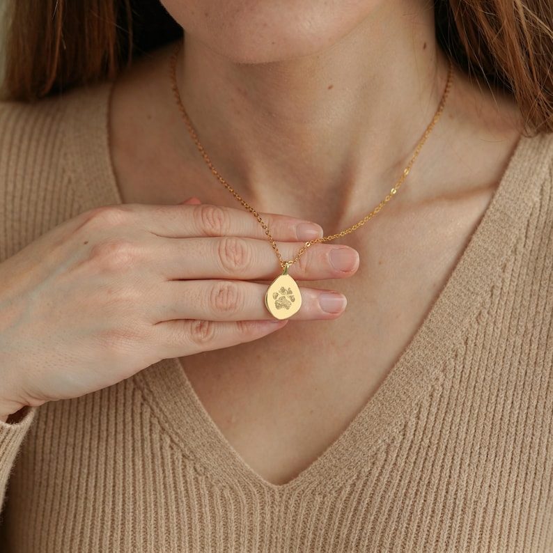 Puede incluir: Un collar de tono dorado con una cadena fina y un peque&ntilde;o colgante de forma irregular similar a una gota. El centro del colgante presenta un grabado detallado de una huella de pata, creando un punto focal sutil y texturizado sobre el metal liso.